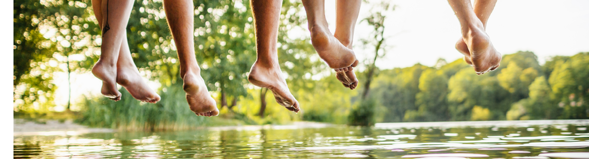 A group of friends legs dangling off a jetty, by the water at a lake together on a sunny afternoon.