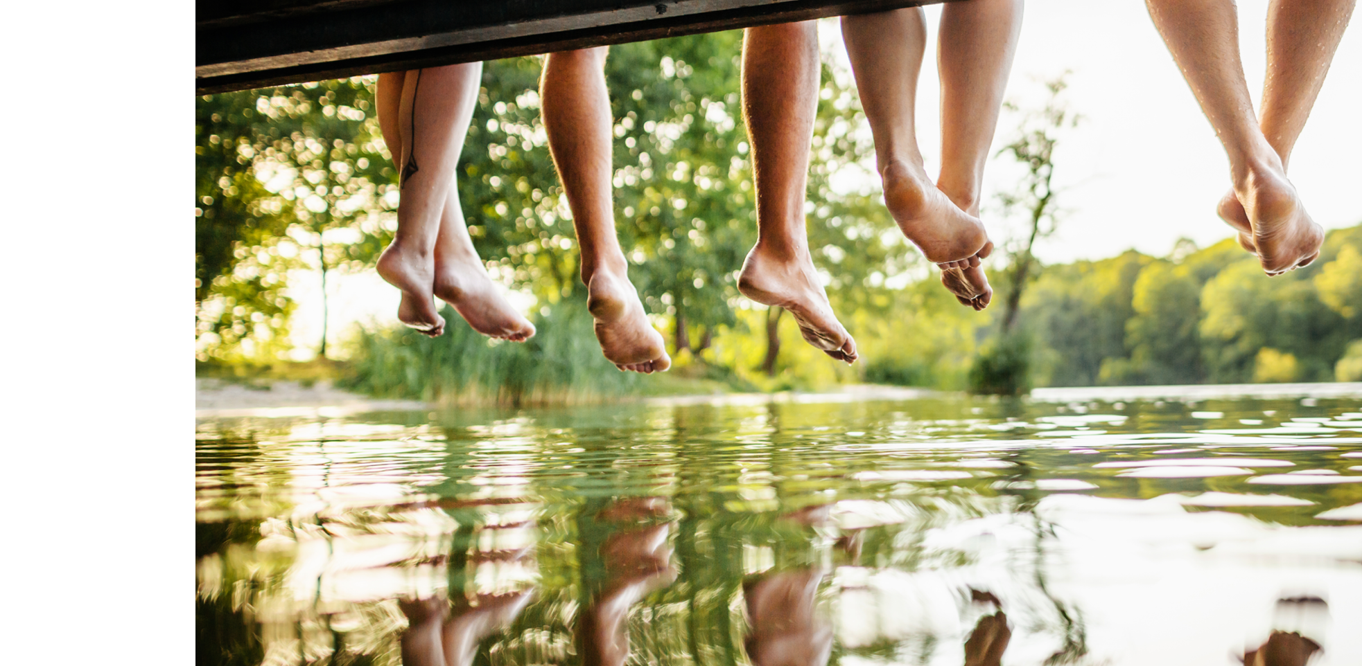 A group of friends legs dangling off a jetty, by the water at a lake together on a sunny afternoon.